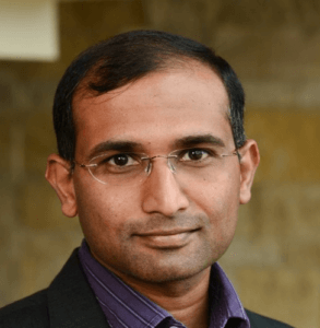 Portrait of Shri. Satish Srinivasan with rimless glasses and a dark blazer on a stone backdrop