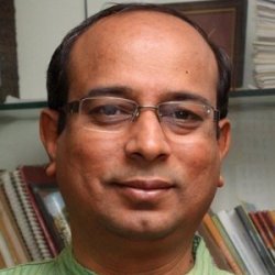 Dr. DK Hari with glasses in a green kurta seated in front of a bookshelf at his office desk