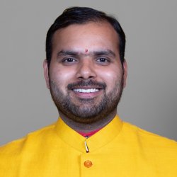 Smiling HUA faculty member wearing a yellow kurta with a red tilak on his forehead, gray backdrop